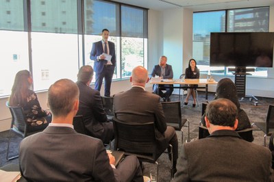 A group of people attending a presentation in an office boardroom. Two presenters are seated at a table at the front of the room. A third person is addressing the audience.